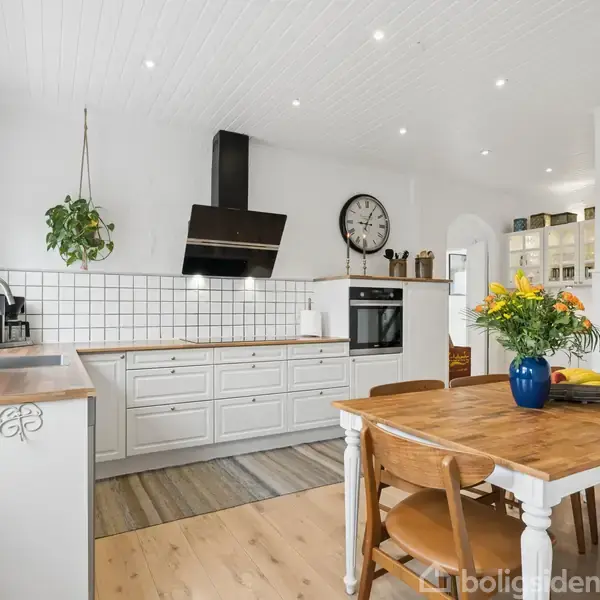 Et gråt hus med skråt tag står i en anlagt have med strukturerede buske og stier. En overdækket terrasse grænser op til huset. Andre huse ses i baggrunden.