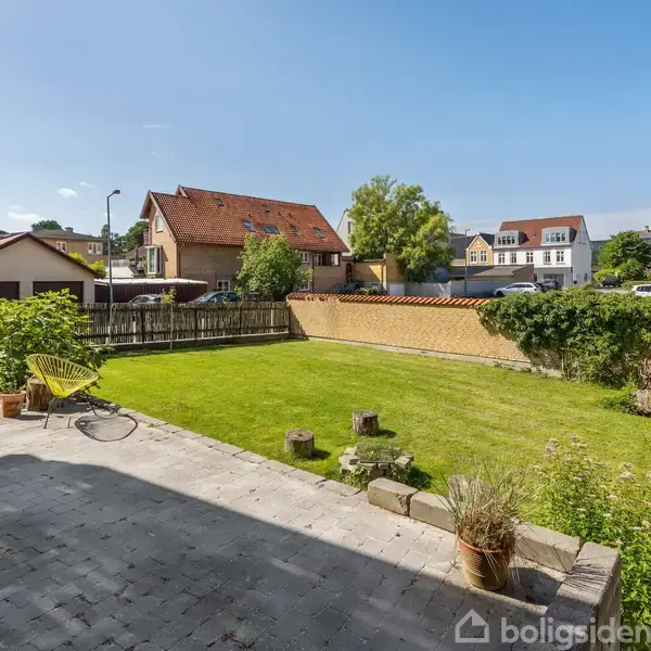 En grøn græsplæne omgivet af et lavt hegn og flisebelagt terrasse. I baggrunden er der huse og et træ, med en gul stol placeret på terrassen.