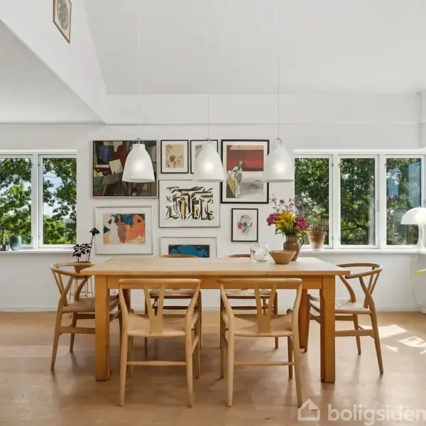 Træbord med stole står i en spisestue, omgivet af store vinduer og kunst på væggen. Hele vægge dækket af glas skabe til højre og blomsterdekorationer på bordet.