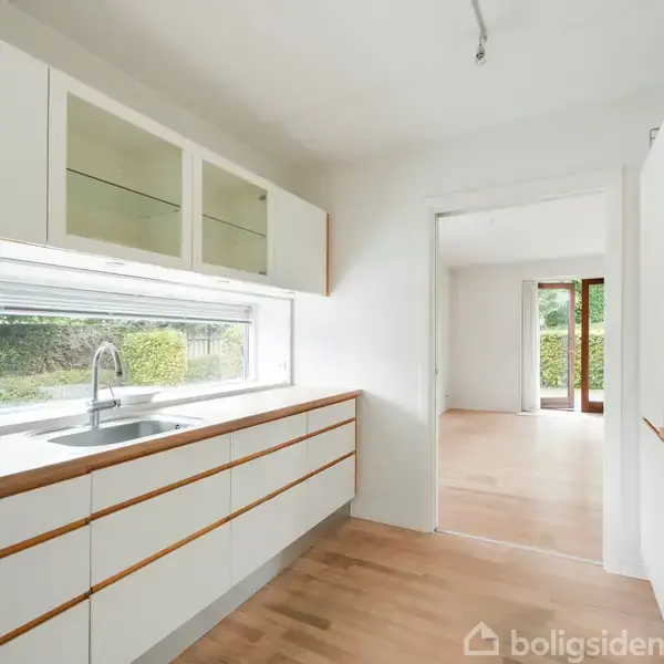 Et moderne køkken med hvide skabe og træbordplader står stille. Store vinduer lader lys ind, mens en dør åbner til en lys stue med trægulve.