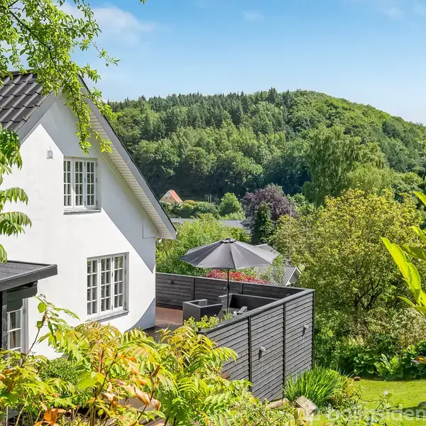 Et hvidt hus med en udendørs terrasse, der har en parasol, omgivet af frodig grønt og træer, placeret i en skovklædt bakkelandskab under en blå himmel.