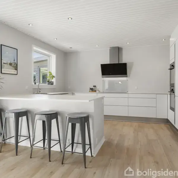 Et moderne køkken har en hvid ø med fire barstole, lyst trægulv og hvide skabe. Væggene er lyse, og der er to vinduer, en emhætte over komfuret, samt potteplanter som pynt.