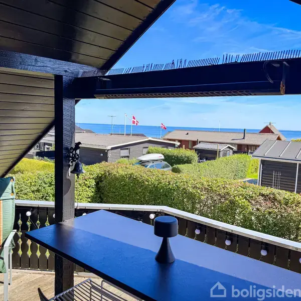 Bord på en overdækket terrasse med udsigt over sommerhuse, havet og to danske flag i baggrunden. Grønne hække afgrænser grunde. Klar blå himmel skaber en solrig atmosfære.