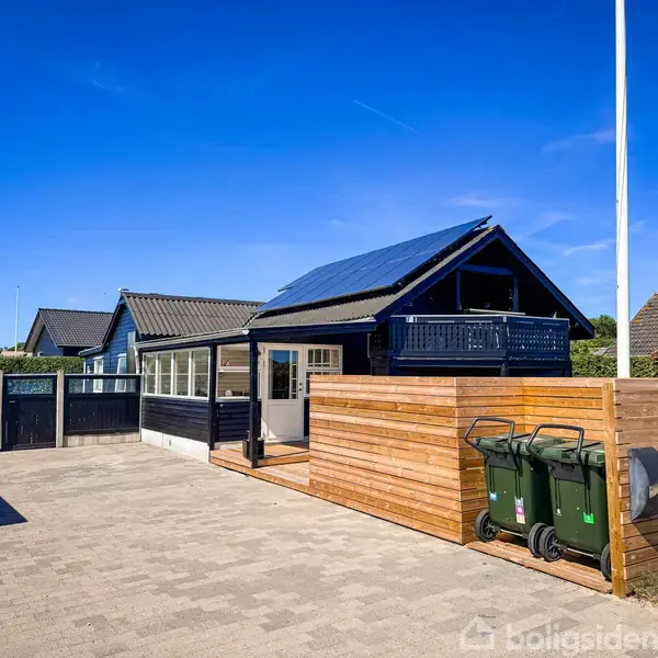 Et moderne, sort hus med solpaneler på taget står ved siden af en træbeklædt skraldespandsskjuler. Omkring ligger andre huse og en indkørsel under en klar blå himmel.