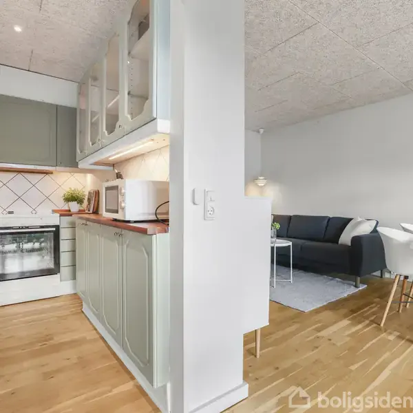 Køkken og stue i åbent plan. Køkkenet har grønne skabe, komfur og mikroovn. I stuen står en mørk sofa ved et lavt bord og en spiseplads med hvidt bord og stole.