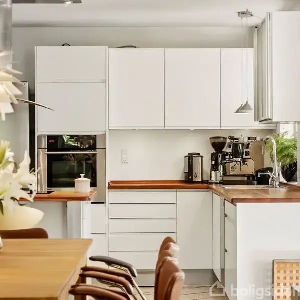 Et moderne køkken har hvide skabe og træbordplader. Til højre står en espressomaskine ved siden af en plante. En træspisebord med blomsterdekorationer står i forgrunden.