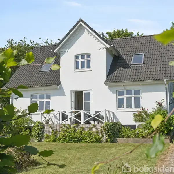 Hus i hvid farve med sort tag står stille omgivet af grøn vegetation i en have. Har en veranda foran og flere vinduer på taget.