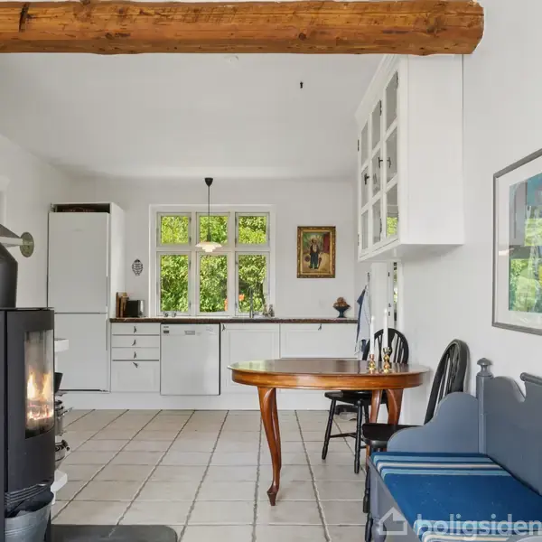 En brændeovn står i en stue med åben udsigt til køkkenet, hvor et rundt bord og blå bænk er placeret. Køkkenet har hvide skabe og et vindue med naturligt lys.