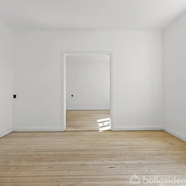 Et tomt rum med hvide vægge og et trægulv, hvor en dør med glasvinduer åbner til et tilstødende rum. Vindue til højre med radiator under. Ingen tekst i billedet.