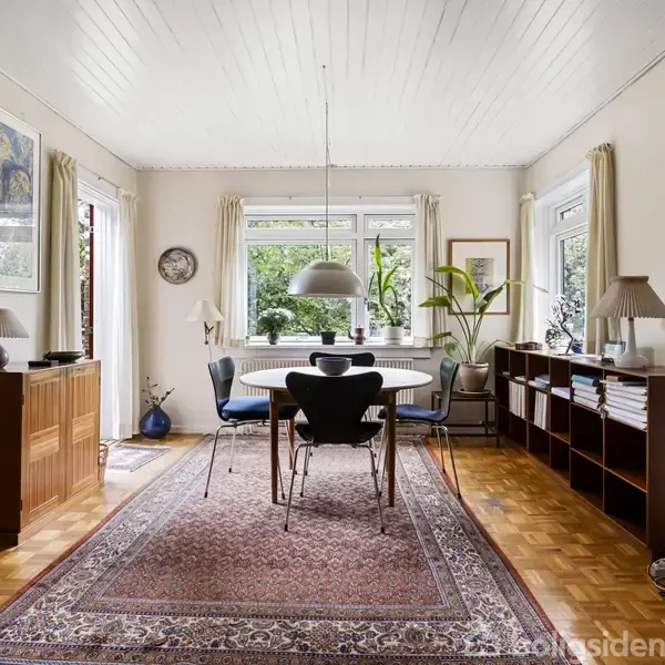 Et spisebord med stole står i et lyst rum med trægulv og tæppe. Væggene har store vinduer, billeder og bogreoler. Planter ved vinduerne skaber en indbydende atmosfære.