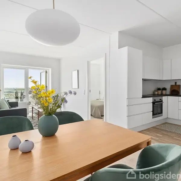 Et spisebord med vase og blomster står i et lyst rum med udsigt til en altan i baggrunden. Et moderne køkken ses til højre.