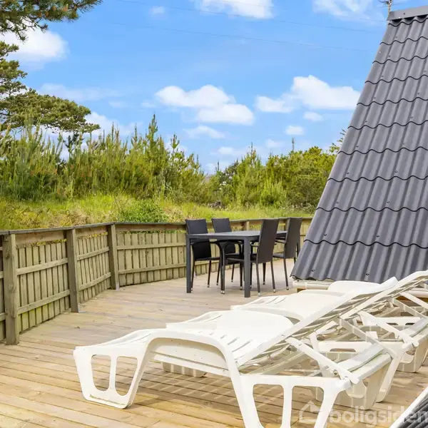 Liggestole står på en træterrasse ved siden af et bord med stole. I baggrunden er der en træhegnet have og træer under en blå himmel.