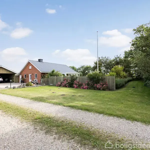 Hus med sort tag ses ved siden af en velholdt have med blomsterbede og græsplæne. Skov og himmel med få skyer i baggrunden.