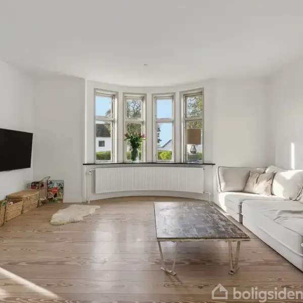 En lys stue med en hvid sofa står omkring et rustikt træbord. Et TV er vægmonteret til venstre. Store vinduer med planter giver udsigt til træer. Kurve står nedenunder.