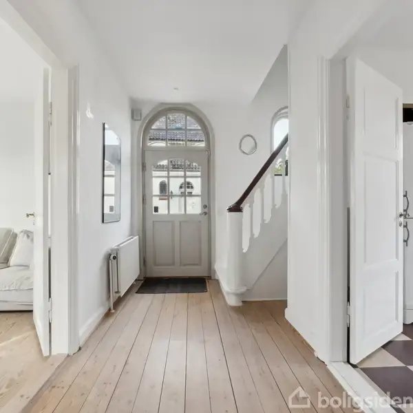 En entré med lyse trægulve og en hvid dør med glasruder fører til en trappe. Til venstre ses et værelse med sofa, og til højre en åben dør.