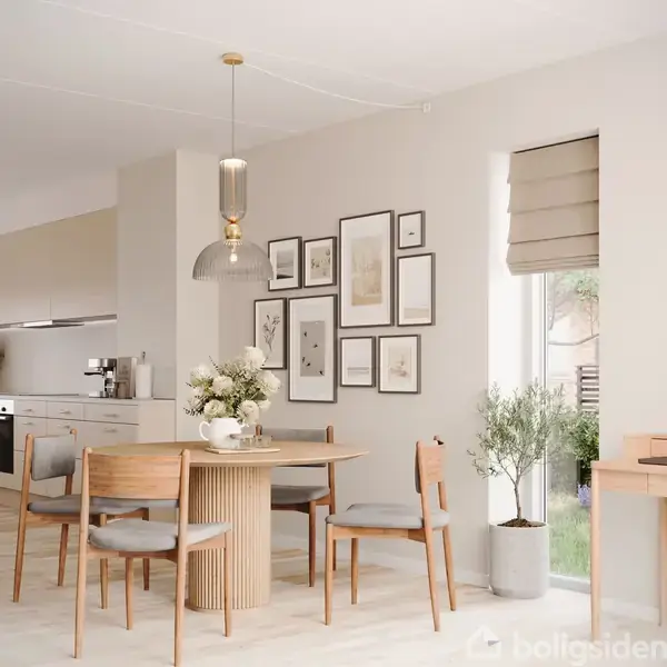 Et spisebord med fire stole står i et lyst køkkenalrum. Vægge er dekoreret med billeder, og store vinduer lukker sollys ind. Der er lyse trægulve og moderne indretning.