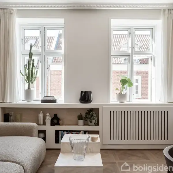 En lys stue med en grå sofa, to store vinduer med hvide gardiner. En kaktus og en anden plante står i vindueskarmen. Hylder under vinduerne har dekorative genstande.