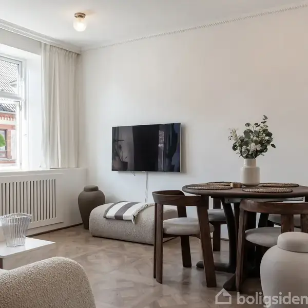 En stue med et rundt spisebord og fire stole. Et fjernsyn hænger på væggen. Der er en sofa, et vindue med lysindfald, og en skammel med en vase på gulvet.