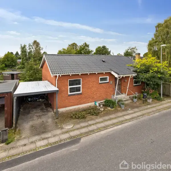 Et rødt murstenshus står stille ved en vej med flisebelægning, omringet af grønne træer og buske. Ved siden af huset er der en carport med cykler.