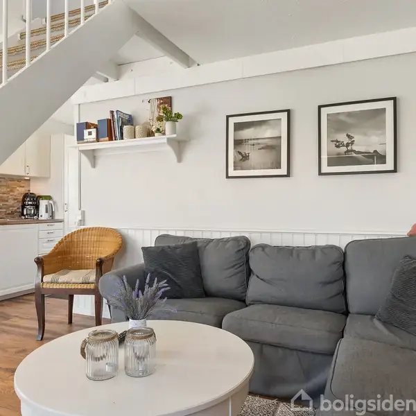 Grå hjørnesofa står i stue med træpaneler, to sort-hvide billeder over sofaen. Rundt sofabord har to lys, ved siden af står en stol. Trætrappe fører op.