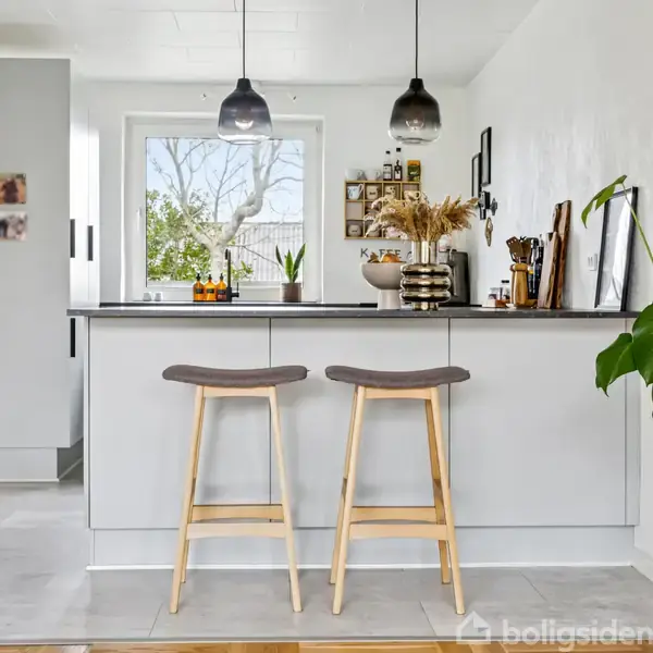 To barstole står ved en køkkenø med grå skabe; ovenfor hænger to sorte lamper. I baggrunden ses et vindue med udsigt til en have, dekorative planter og køkkenudstyr.