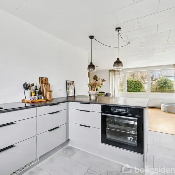 Køkken med hvide skabe og sorte bordplader; der står køkkenredskaber på bordet. I baggrunden ses en stue med store vinduer og grøn udsigt. Lyse lamper hænger fra loftet.