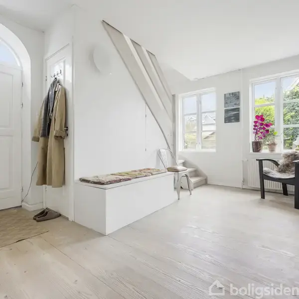 En lys entré med en hvid dør står åben, frakker hænger på væggen, en bænk med farvet pude står ved siden af. Vinduer viser grønt udenfor, og en stol med et bord ses nær dem. En plante står på bordet.