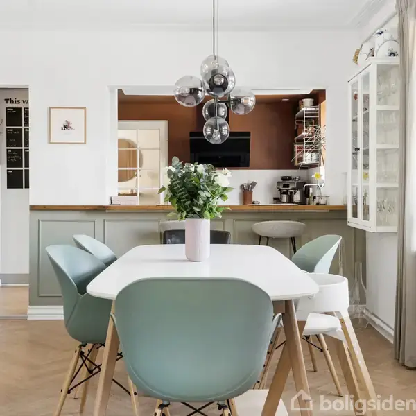 Et moderne spisebord med lyseblå stole står i et lyst rum med trægulve. Der hænger en stilfuld lampe ovenover, og i baggrunden ses et køkken med barstole og planter.