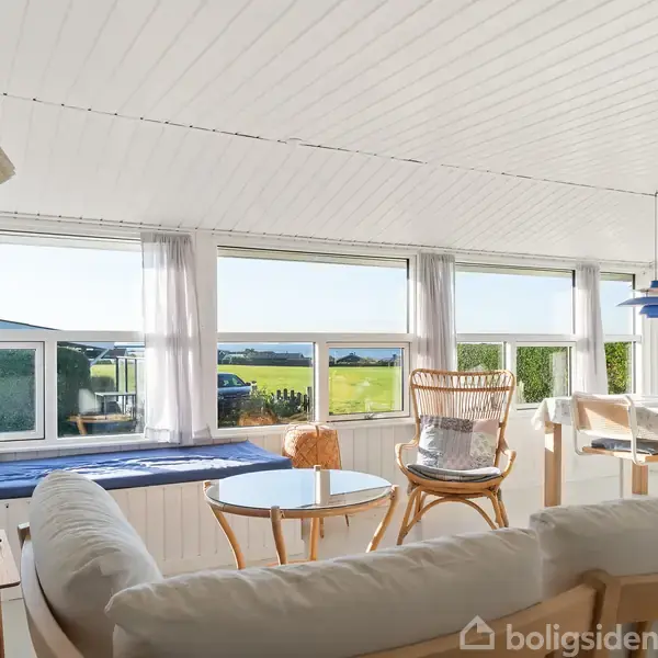 En stue med hvide vægge og træmøbler har en sofa i forgrunden. Store vinduer indrammer en udsigt til grønne marker og blå himmel, mens lamper hænger fra loftet.