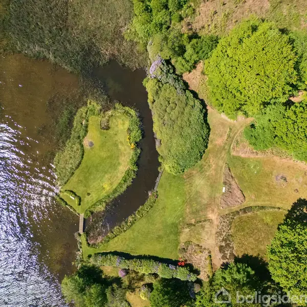 En grøn halvø strækker sig ud i en sø, omgivet af frodige træer. En lille bro forbinder halvøen med bredden. Vandet skvulper blidt omkring et naturligt landskab.