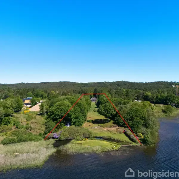 Grønt område langs en sø markeret med en rød trekant, omgivet af frodige træer og buske. I baggrunden ses en skovklædt bakke under en lyseblå himmel.