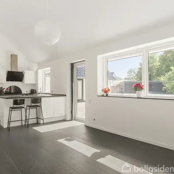 Et lyst køkken med en central ø og to høje stole, omgivet af hvide vægge og mørkt gulv. Store vinduer med udsigt til en have og dekoreret med blomster.