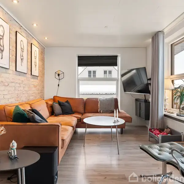 Brun sofa står i hyggelig stue med træbjælkevæg. Runde borde og moderne læderstol ved siden af stort vindue. Tre billeder hænger på væggen. Fjernsyn hænger til højre.