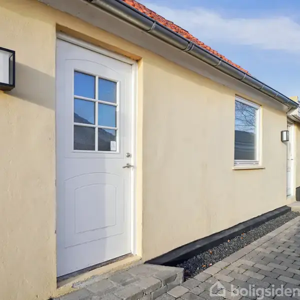 En hvid hoveddør med rammer og rude står i en gul murstensfacade. Bygningen er omgivet af en flisebelagt gangsti, og himlen er klar med enkelte skyer.