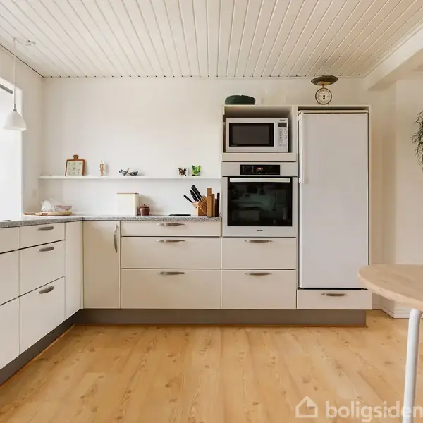 Køkken med hvide skabe og træbordplade. En mikroovn og ovn er indbygget i skabsvæggen. Rummet er lyst med trægulv og hylder med dekorationer.