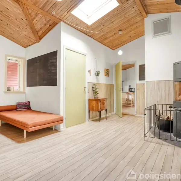 En moderne brændeovn står i et lyst rum med trægulv og loft. Til venstre ses en orange daybed, mens døre og et lille skab omkranser midten af rummet.