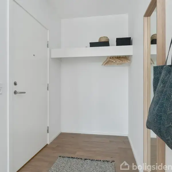 Dør står lukket i en lys entré. En taske hænger på en knage ved spejlet, og flere bøjler hænger på en hylde i baggrunden.