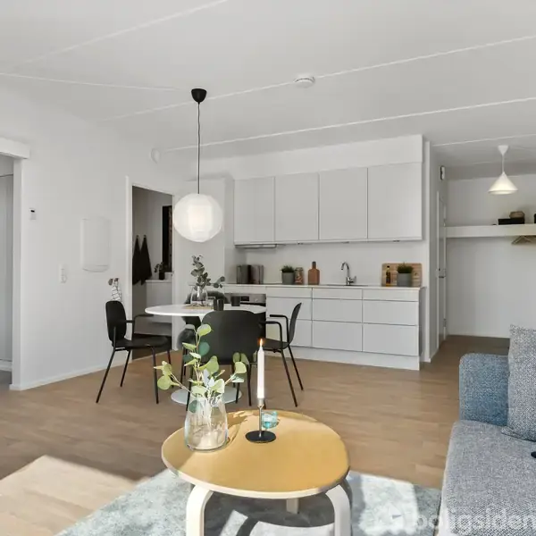 En moderne stue med gråt sofaarrangement, rundt sofabord med planter, og hvidt køkken i baggrunden. Fire stole omringer spisebord, og en pendellampe hænger over bordet.