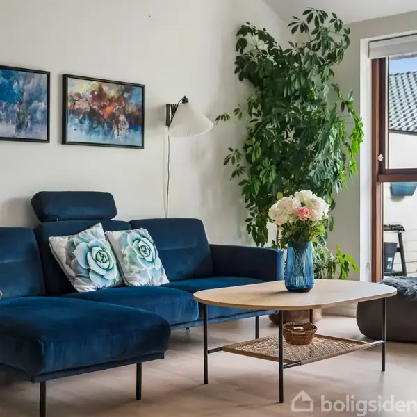 Blåt sofaarrangement med puder står mod en væg med to malerier. En stor grøn plante står ved siden af, mens et rundt bord med en vase med blomster står i midten.