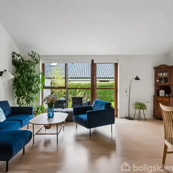 Blå sofa og lænestole står i en lys stue med træmøbler. Store vinduer viser en grøn have udenfor. På væggen hænger billeder og en gulvlampe står ved siden af sofaen. En spisebord med træstole ses til højre.