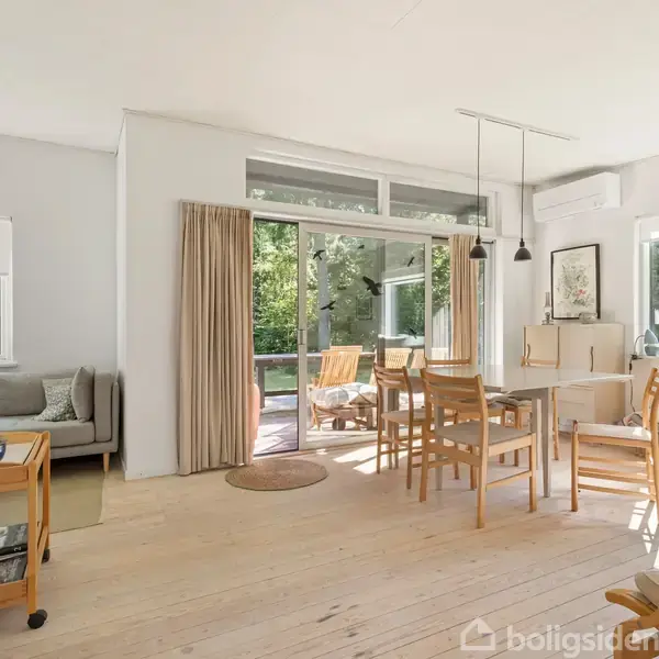 Stue med træmøbler, spisebord og stole, lys indstrømmer gennem store vinduer. En sofa står i hjørnet, og der er adgang til terrasse gennem glasdøre. Nogle malerier hænger på væggene.