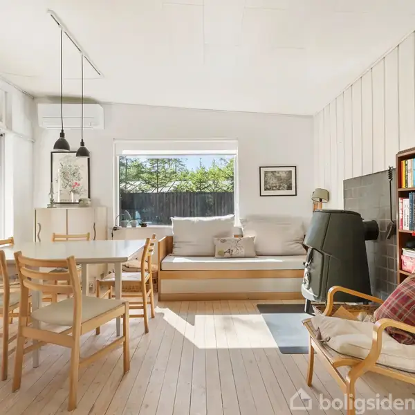 En lys stue med et spisebord og stole i træ, en daybed under vinduet, en pejs ved siden af en bogreol fyldt med bøger og en lænestol.
