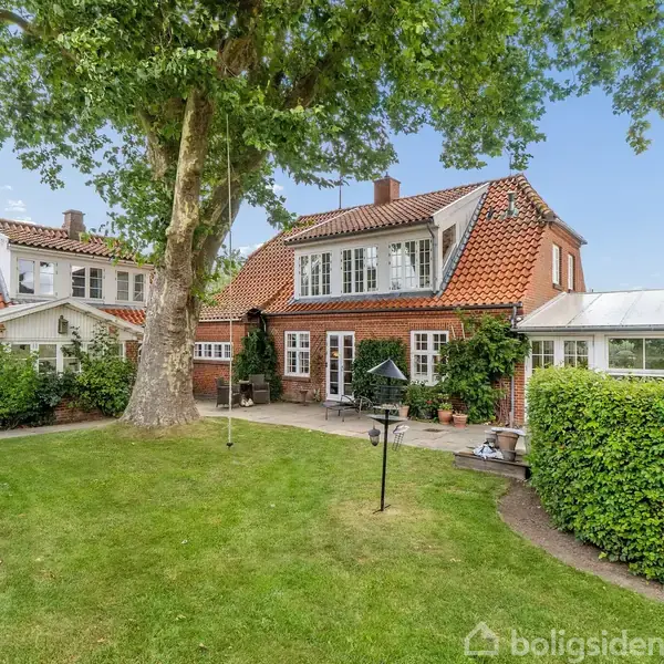 Et rødt murstenshus med rødt tegltag står i en grøn have med en stor træ ved siden af. Sollys skinner på huset, omgivet af buske og græs.