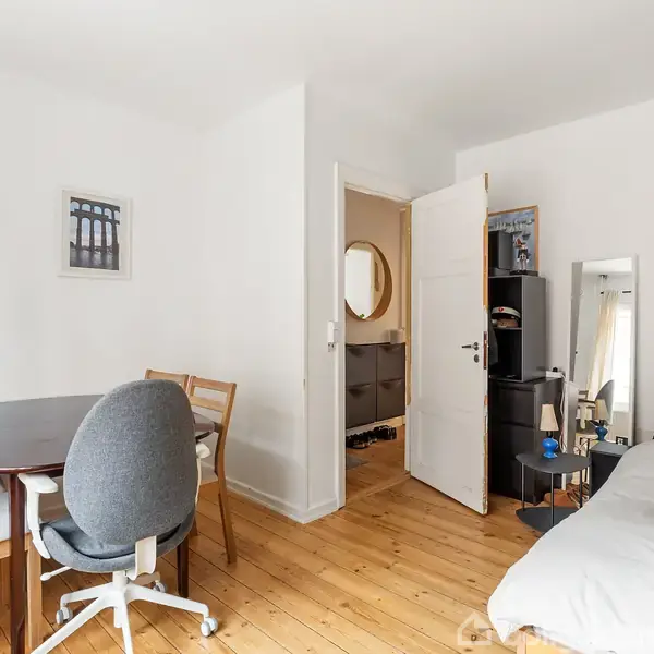 Et lyst værelse med trægulv indeholder et bord og stole i venstre side, en grå kontorstol, en seng til højre, billeder på væggen og en åben dør. En stå-lampe og reol er i baggrunden.