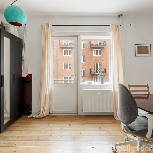 Et værelse med et mørkt klædeskab, en drejestol ved et rundt bord, og en dør til en altan. Væggene er hvide, og der hænger en blå lampe.
