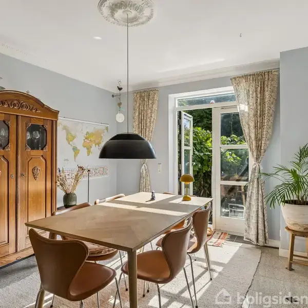 Et spisebord med stole står midt i et lyst rum. En lampe hænger over bordet. Store vinduer med gardiner åbner ud til en have. En træskænk står ved væggen.