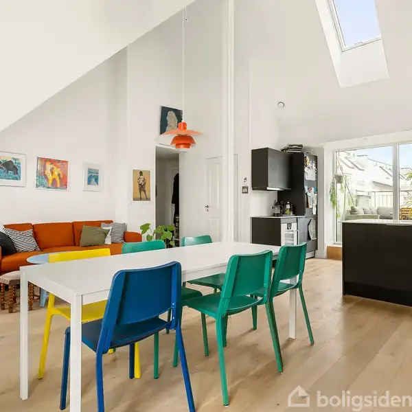 Et spisebord med farverige stole står i et lyst, moderne rum. Ved siden af er en orange sofa med puder. Store vinduer bagved giver lys og udsigt til planter udenfor.