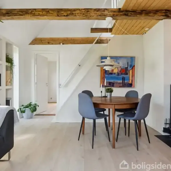 Rundt spisebord med fire stole står i en lys stue med træbjælker i loftet. En tændt brændeovn til højre skaber hyggelig atmosfære. Møblering inkluderer også reol og sofa.