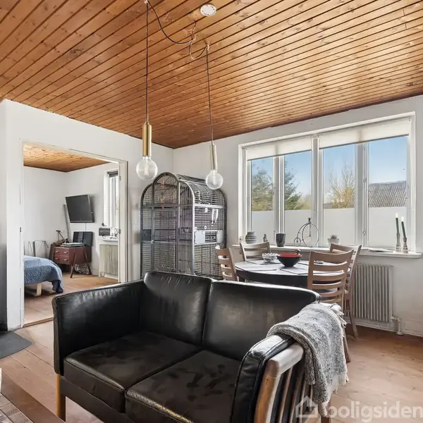En sort sofa står i stuen, ved siden af et spisebord foran et vindue. Der er en brændeovn til venstre og træloft, med en åben dør til et soveværelse.