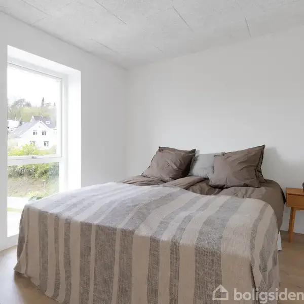 En dobbeltseng dækket med et stribet tæppe står i et lyst soveværelse. Ved siden af sengen er et lille trænatbord med en lampe og bøger.